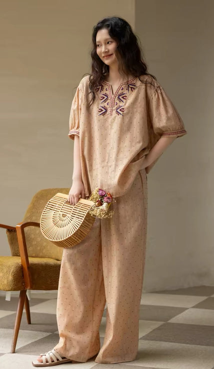 Woman in a beige embroidered outfit holding a woven bag in a room with a chair and decorative items.