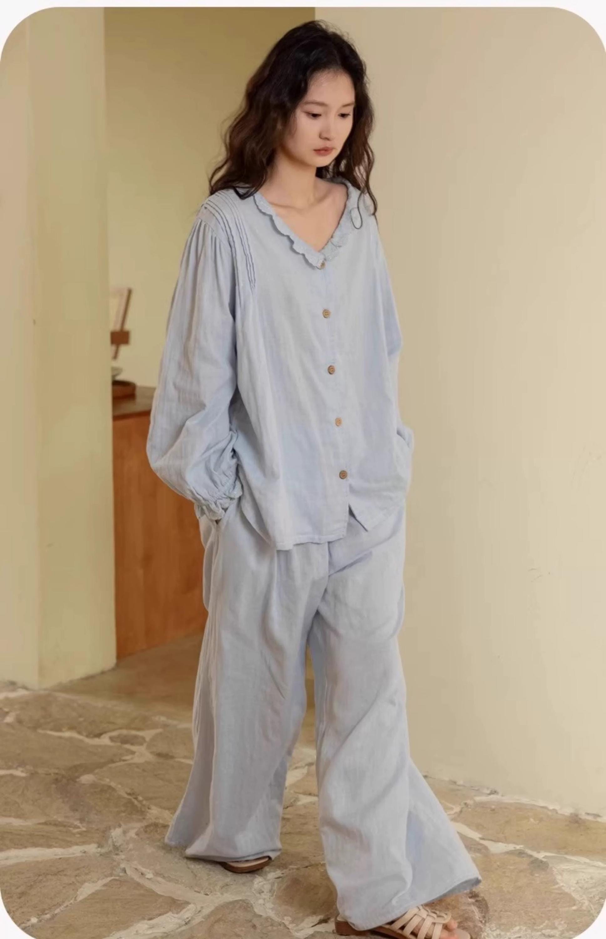May include: A light blue pajama set. The top has a ruffled collar and button-up front. The long sleeves have gathered cuffs. The pants are wide-legged. The fabric appears soft and lightweight. The outfit is displayed on a person standing in a room with a neutral-colored wall